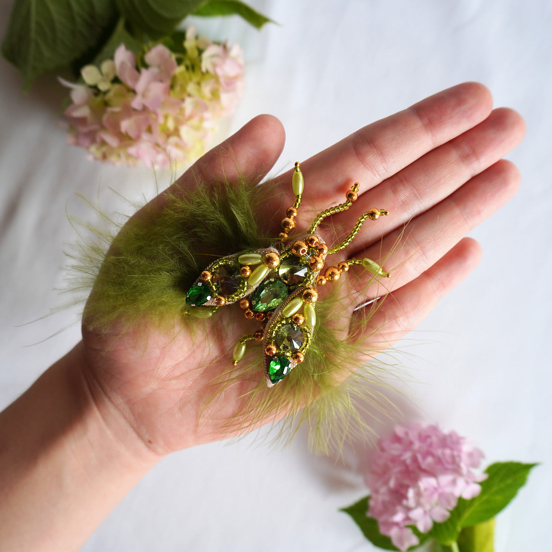 Hand holding a decorative fly TGbrooch with green stones and gold accents